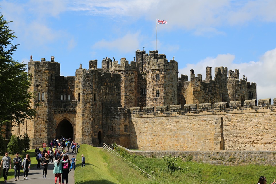 Visitors walking towards the grand entrance of a medieval stone castle under a clear sky, with the uk flag flying atop one of the towers.