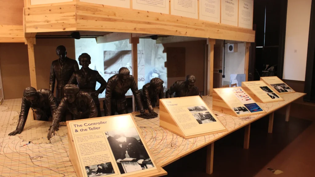 A museum exhibit featuring bronze statues of men in various poses, and display boards with text and images, all set on a map-covered floor under a wooden platform.