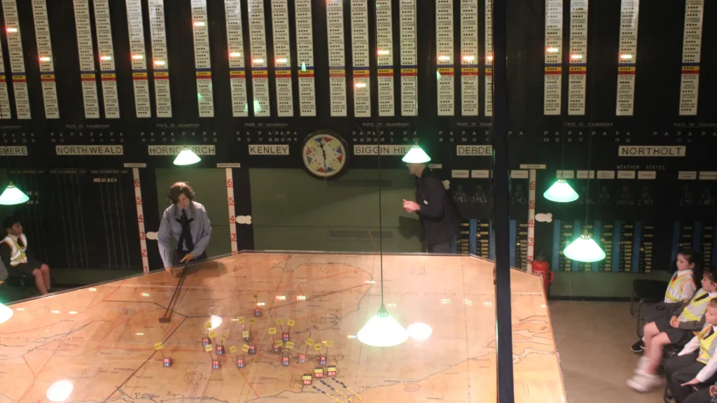 Inside a historical war room, visitors explore a large map table with colored markers under overhead green lamps, surrounded by wall panels displaying various location names.