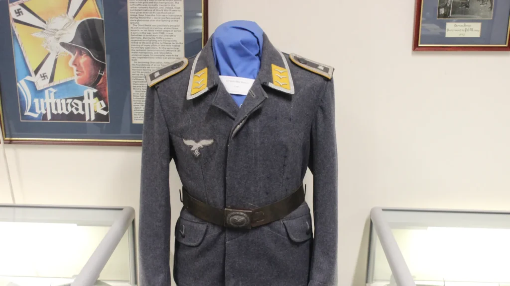 A world war ii luftwaffe uniform displayed on a mannequin, featuring insignias and a belt, exhibited in a museum with historical posters in the background.