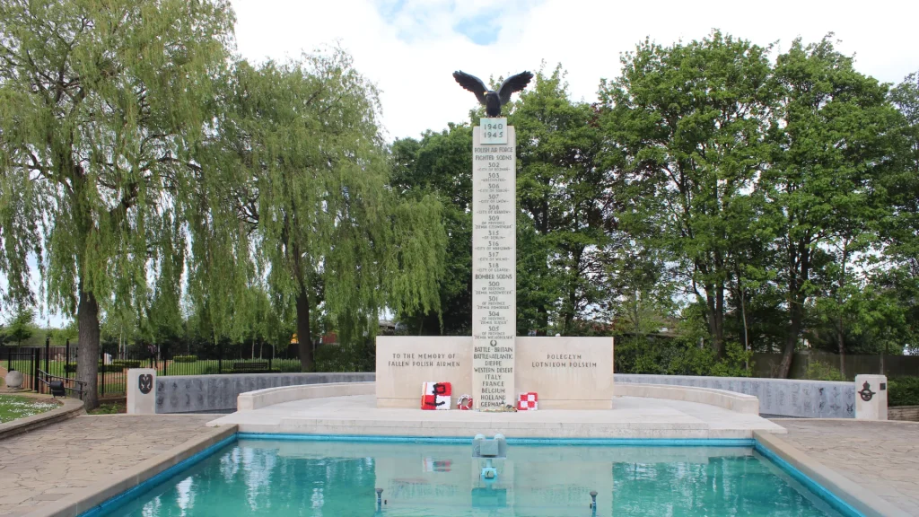 A war memorial with a vertical column topped by an eagle, flanked by weeping willow trees and fronted by a reflective pool, against a cloudy sky.