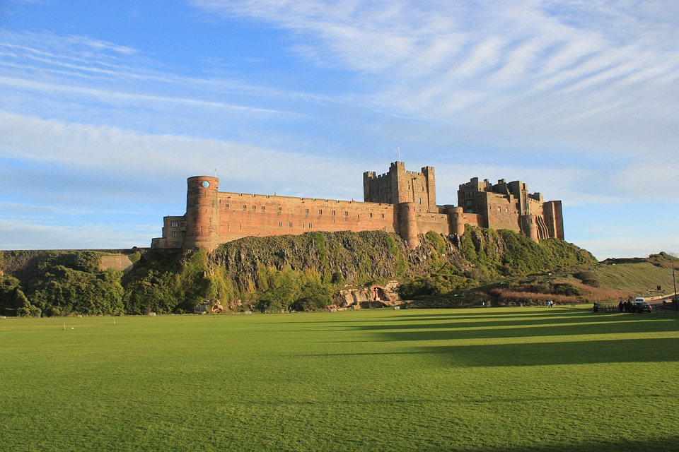 An expansive view of a large medieval castle with multiple towers and sections, perched on a rugged hill, illuminated by sunlight against a backdrop of blue skies and fluffy clouds, with a vast green lawn in the foreground.
