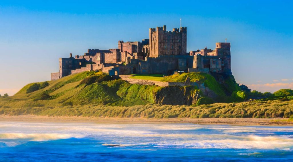 A majestic ancient castle perched atop a lush green hillside near a sandy beach with waves gently crashing in the foreground under a clear blue sky.