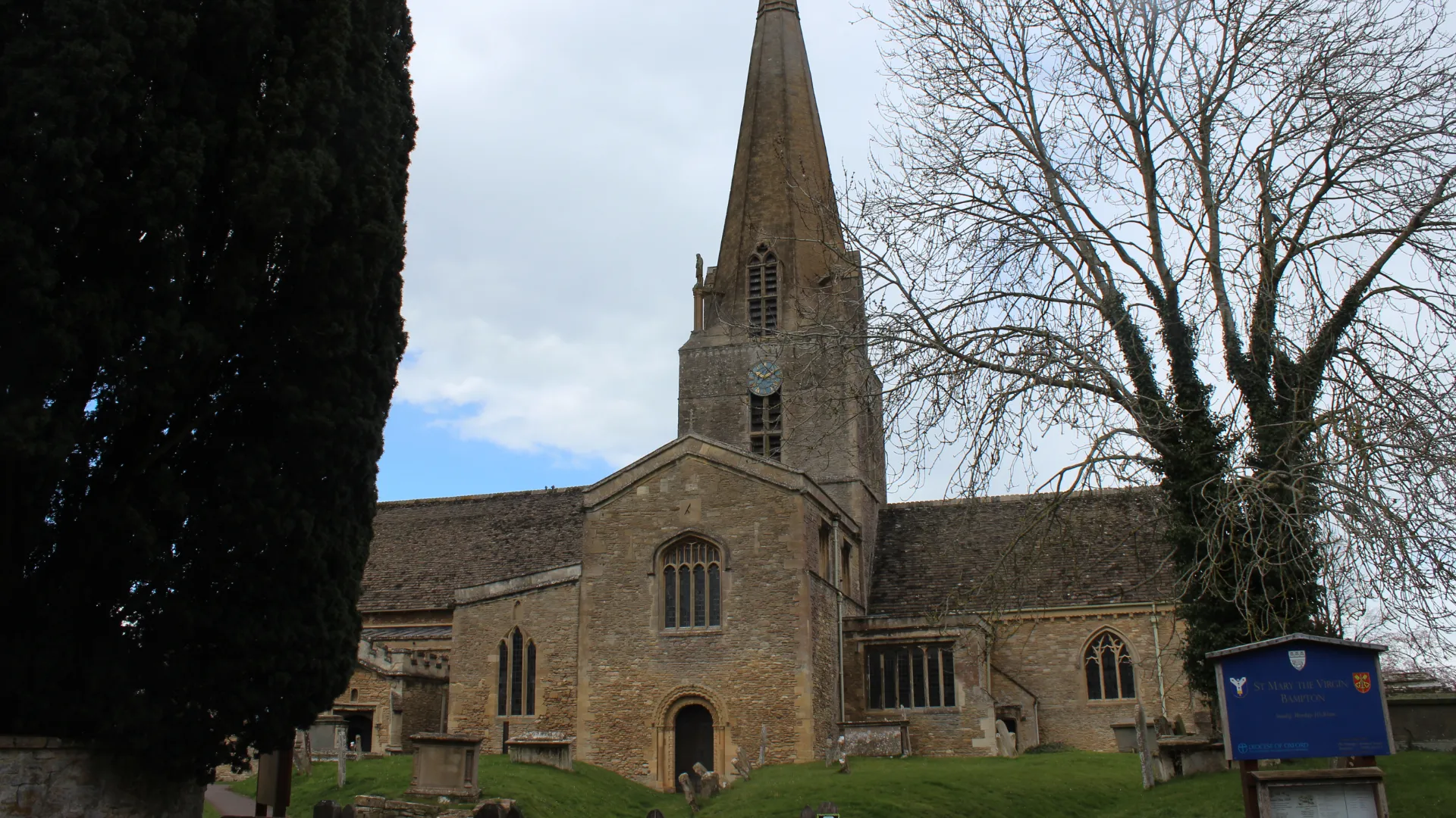 A historic stone church with a tall, pointed spire, surrounded by leafless trees under a cloudy sky. a blue information sign is visible near the entrance.