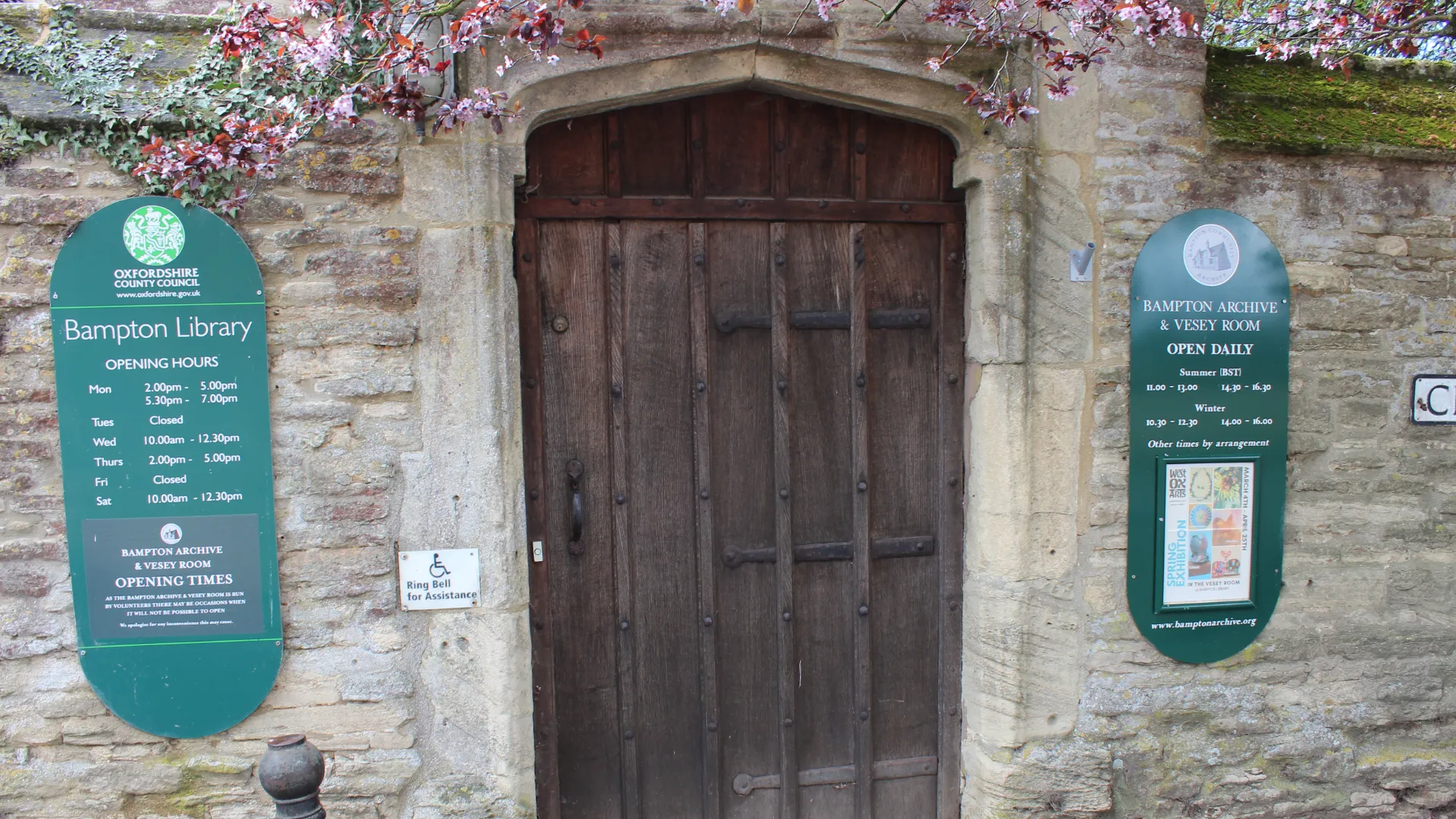 Old wooden door flanked by two informational signs about bampton library and archive, displayed on a stone wall with blooming branches above.