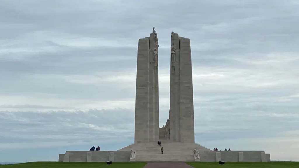 The canadian national vimy memorial stands under a cloudy sky, featuring twin limestone towers with statues, honoring fallen soldiers. visitors are seen walking around the monument.