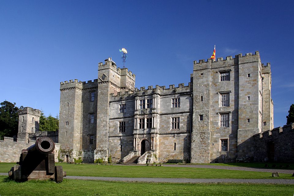 A large, historic stone castle under a clear blue sky, featuring a prominent central building with a grand entrance and multiple towers, flanked by an old cannon on the lawn in the foreground. flags fly atop the towers.