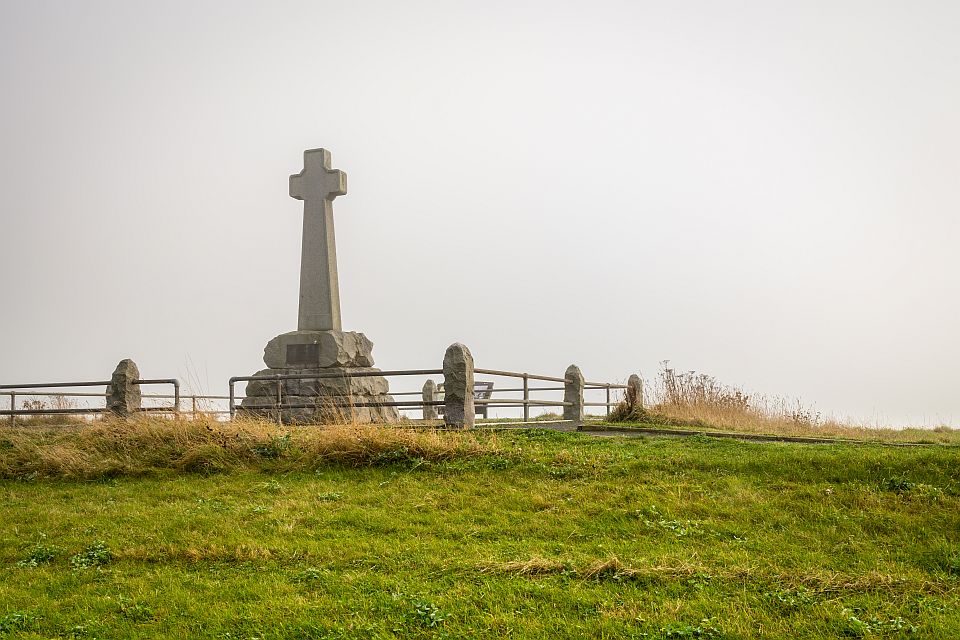 A large stone cross monument stands on a grassy hill surrounded by smaller stone posts, with a misty backdrop.