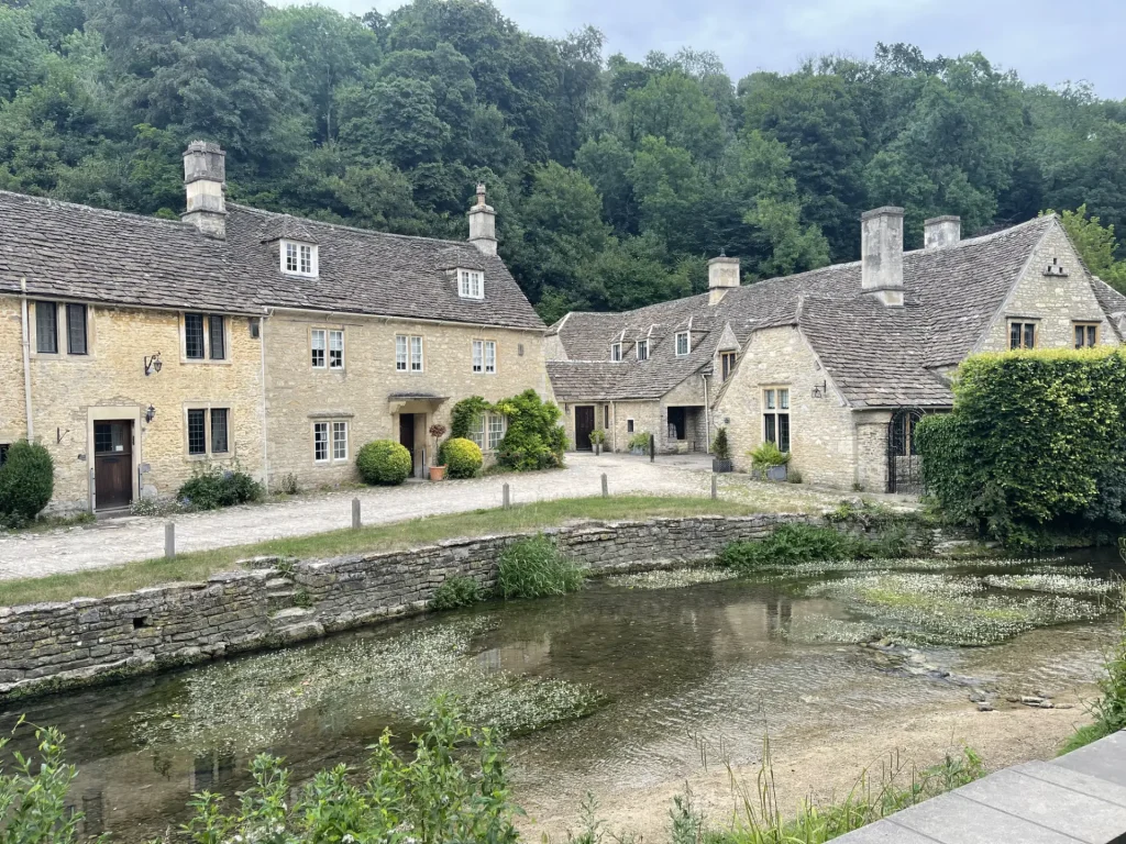 Charming view of a quaint cotswold village featuring traditional limestone houses alongside a serene stream, surrounded by lush greenery.