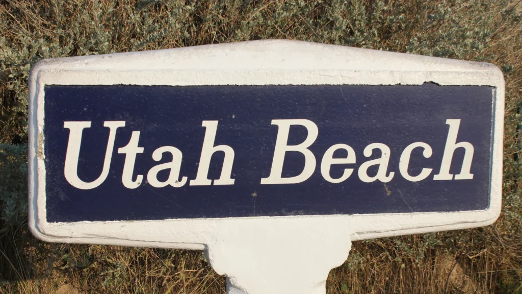 White and blue sign reading "utah beach" set against a backdrop of sandy and grassy terrain.