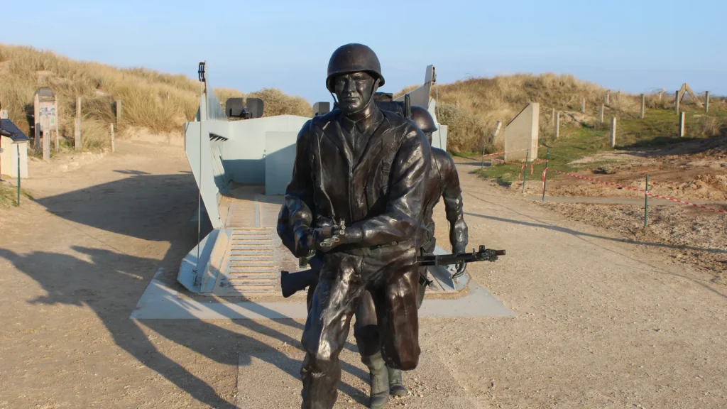 A bronze statue of a soldier in full military gear holding a rifle, standing in front of a beach landscape with sand dunes under a clear sky.