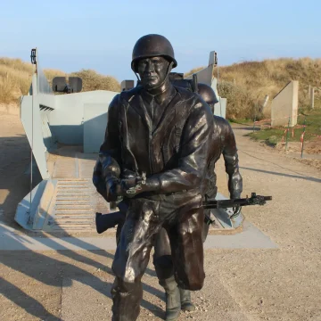 A bronze statue of a soldier in full military gear holding a rifle, standing in front of a beach landscape with sand dunes under a clear sky.