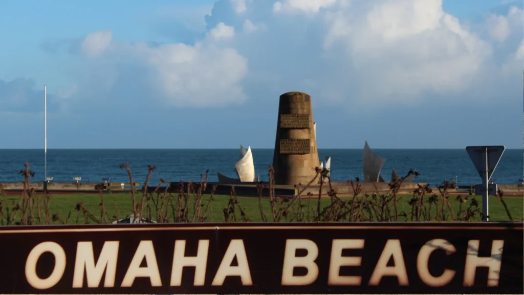 View of omaha beach monument with "omaha beach" sign in foreground. the monument stands against a clear sky, contrasting with the serene blue sea in the background.