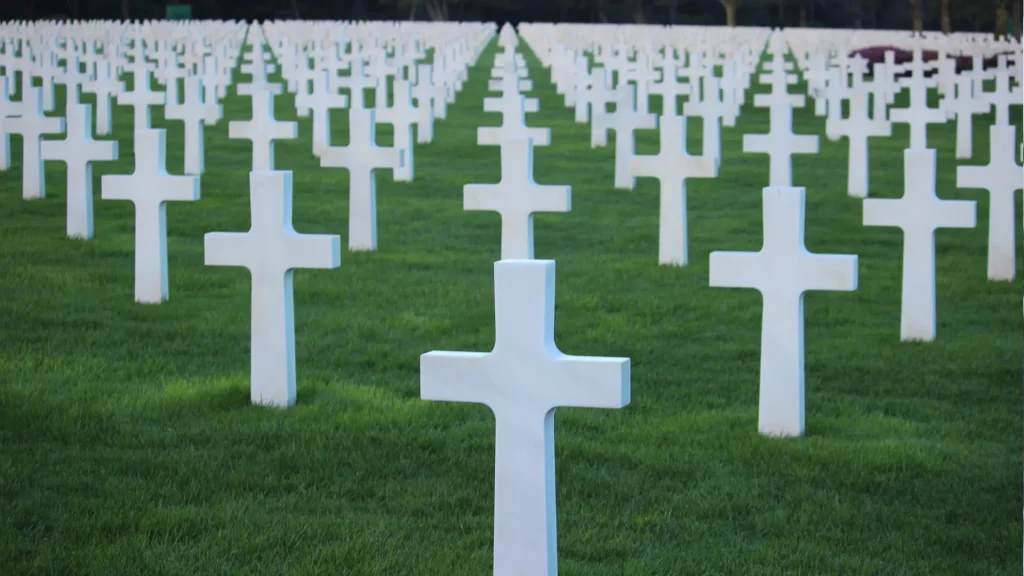 A view of countless white crosses arranged in rows on a grassy field, symbolizing a military cemetery. the focus is on one cross in the foreground, with others fading into the background.
