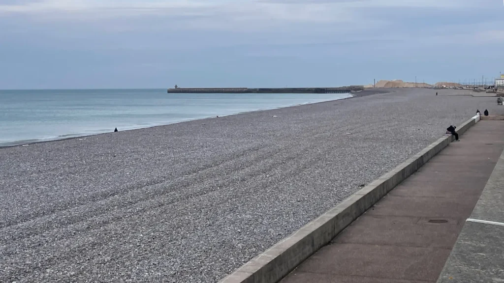 A pebbly beach extends along a calm sea, with a pier in the distance and a clear sky above. few people stroll along the adjacent promenade.