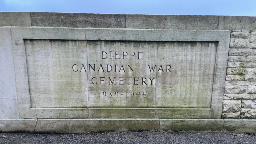 Alt text: a weathered stone plaque at the entrance of the dieppe canadian war cemetery, inscribed with "dieppe canadian war cemetery 1939-1945" on a solid wall.