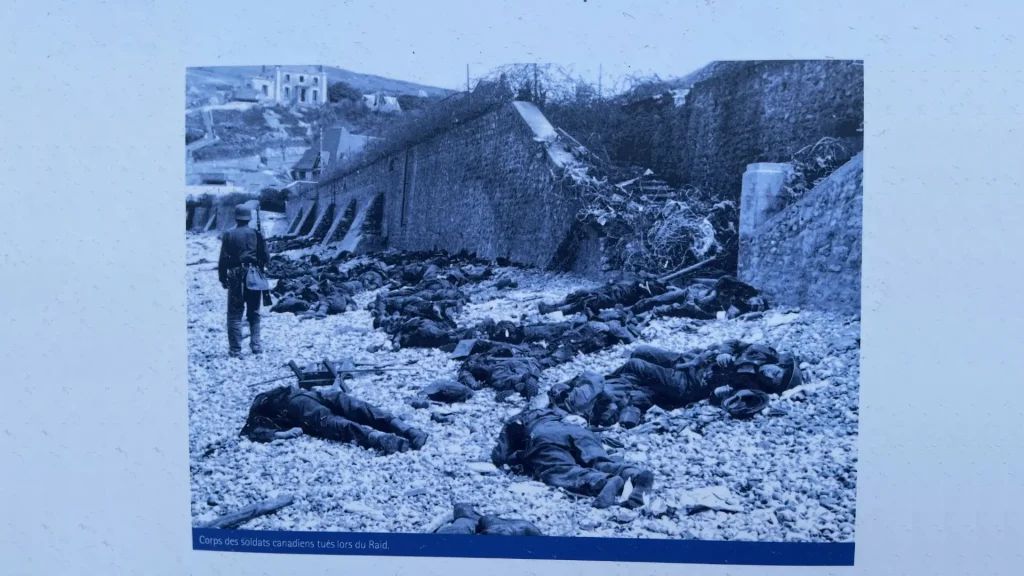 A historical black-and-white photo showing a group of soldiers lying on the ground, presumably casualties of war, with a standing soldier observing the scene and buildings in the background.