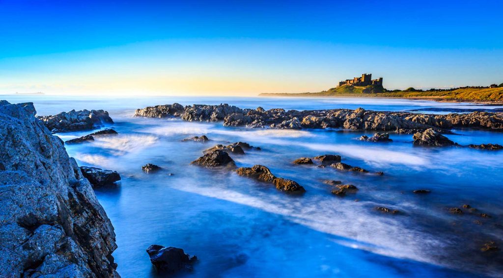 Long exposure shot of a serene coastline with rugged rocks in the foreground, misty ocean water and a castle overlooking the sea in the background under a clear blue sky.
