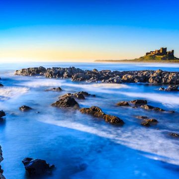Long exposure shot of a serene coastline with rugged rocks in the foreground, misty ocean water and a castle overlooking the sea in the background under a clear blue sky.