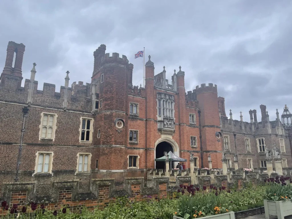 A historical red-brick castle with tudor-style architecture under a cloudy sky, featuring a prominent gatehouse, ornate chimneys, and a vibrant garden in the foreground.
