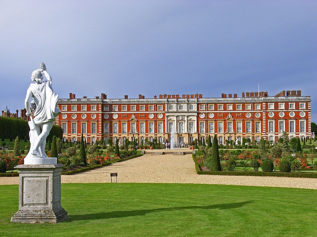 A statue in the foreground overlooking the symmetrical gardens and the expansive hampton court palace with a lush green lawn and cloudy sky.