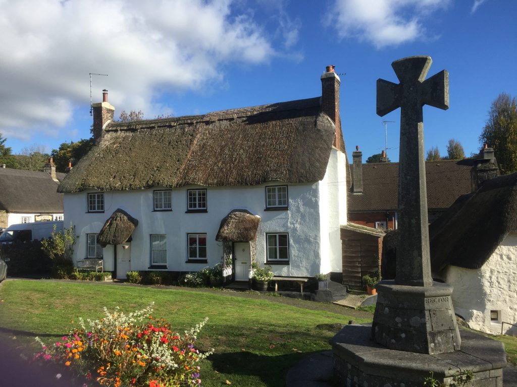 A charming, white, thatched-roof cottage with bright flowers in the foreground and a stone cross on the right, set against a backdrop of other houses under a clear blue sky.