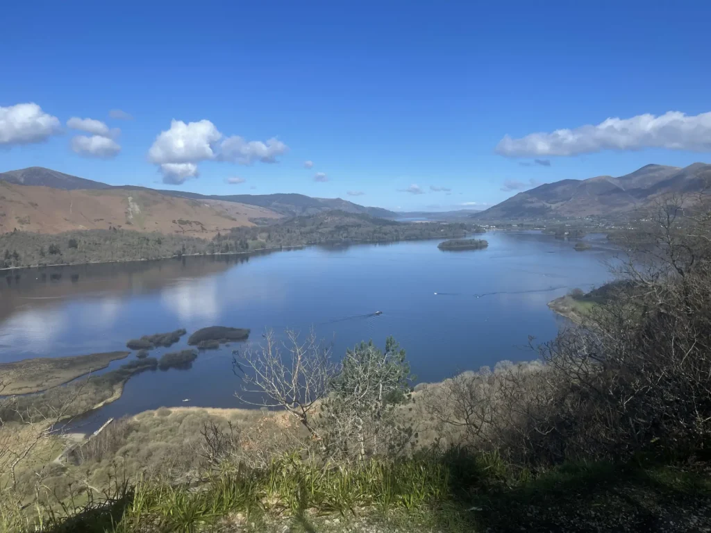 A panoramic view of a serene lake surrounded by rolling hills and sparse vegetation under a clear blue sky with scattered clouds.