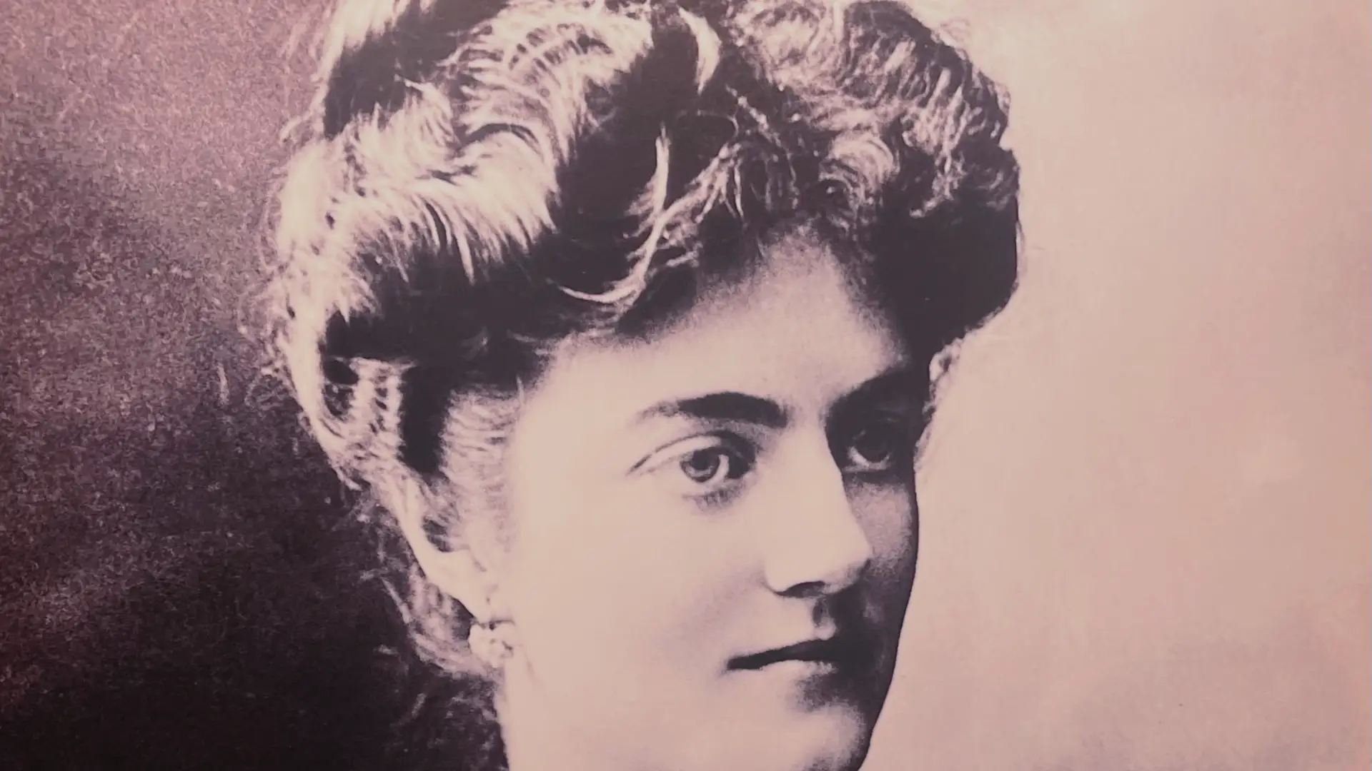 Vintage black and white portrait of a woman with elaborately styled hair, looking to the side, captured in a soft, grainy texture suggestive of early photographic techniques.