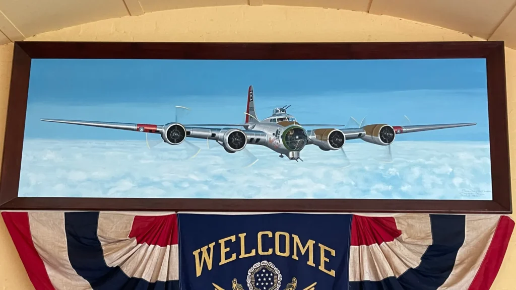 A painting of a vintage four-engine propeller airplane, framed, positioned above a decorative banner with "welcome" on it and a ceremonial backdrop featuring the u.s. flag colors and emblem.