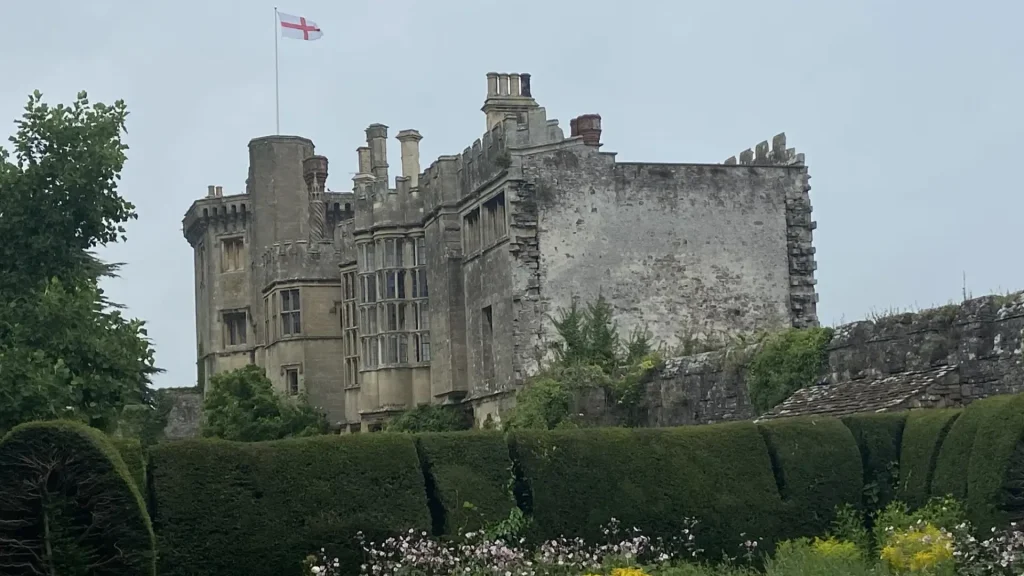 Large, partially weathered castle with a flagpole flying the flag of england, surrounded by lush green hedges and trees under an overcast sky.