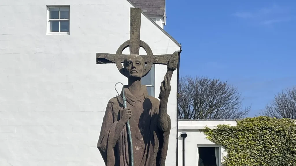 Statue of a religious figure holding a crucifix and staff, standing in front of a white building with a clear blue sky and green foliage in the foreground.
