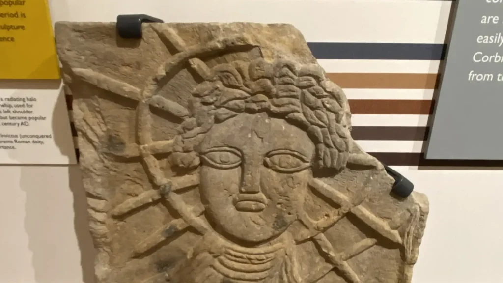 Ancient carved stone relief featuring a detailed bust of a person with radiant lines suggesting an ornate headdress or halo, displayed in a museum.