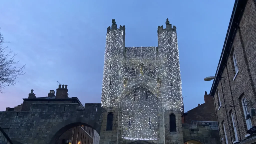 A historic stone gate tower decorated with twinkling string lights against a dusk sky.