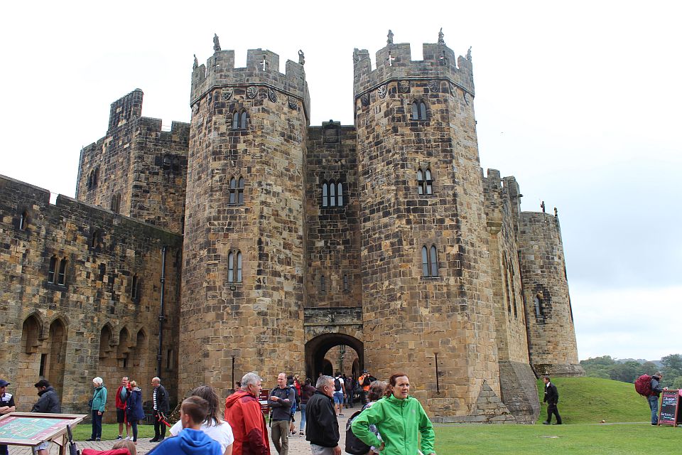 Visitors exploring the grounds of a grand medieval castle with robust twin towers and fortified walls under a cloudy sky.