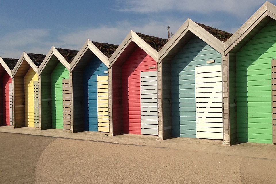 A row of colorful beach huts with peaked roofs in a spectrum of colors including blue, red, yellow, and green, standing under a clear blue sky.