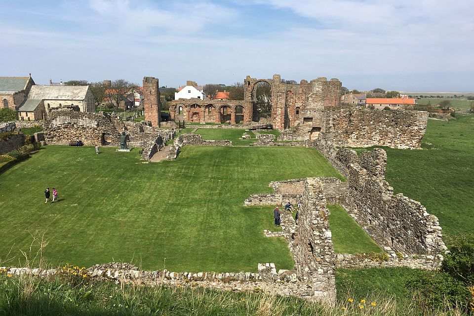 Ruins of an ancient abbey with partially standing walls and archways, surrounded by green lawns. visitors are walking through the site on a sunny day with clear skies.