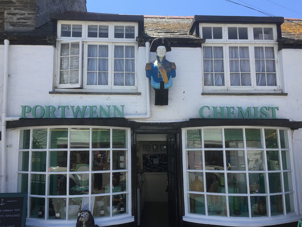A quaint two-story building with a sign reading "portwenn chemist." the facade features a painted character above the name and traditional green-framed shop windows on the ground floor.