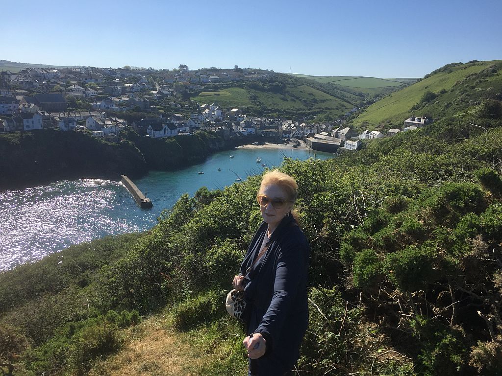A woman in sunglasses stands on a hill overlooking a scenic coastal village with sparkling blue water, visible under bright sunlight.