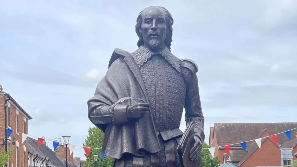 Statue of william shakespeare standing in a traditional elizabethan outfit, set against a sky with light clouds and surrounded by buildings with festive flags.