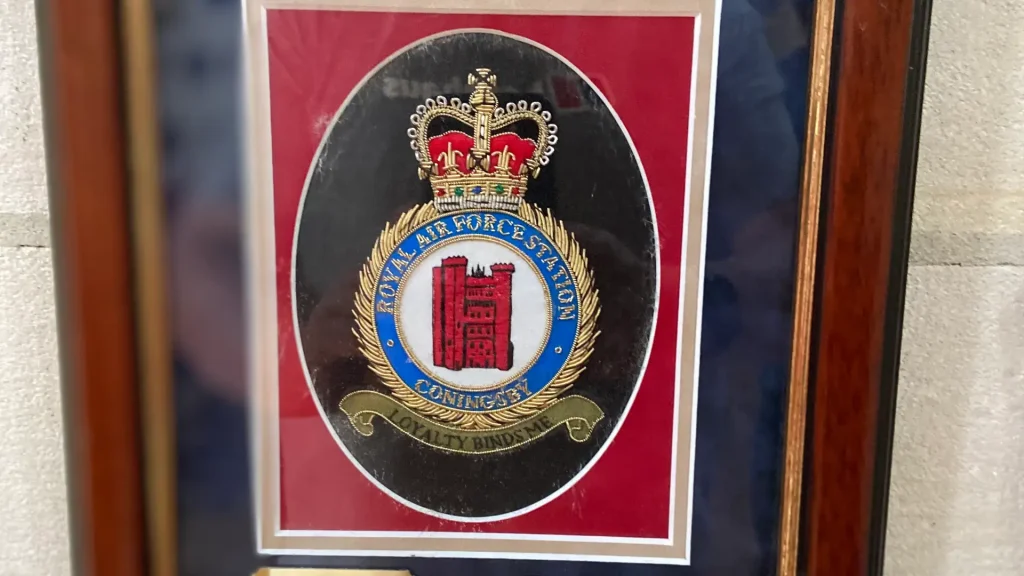 An emblem of the royal air force station coningsby, featuring a red and white castle motif with golden crowns, encircled by a blue band with golden text. it's framed and displayed on a wall.