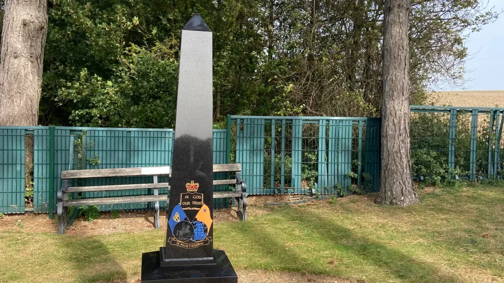 A black obelisk-shaped memorial with emblems and text stands in a park area, flanked by a wooden bench and surrounded by trees and a green fence.