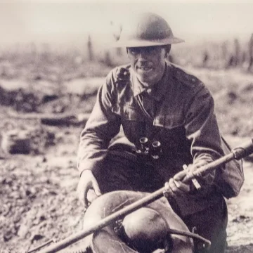 Black and white historical photo of a world war i soldier sitting on the battlefield with a rifle and helmet, smiling at the camera, amidst a desolate landscape.