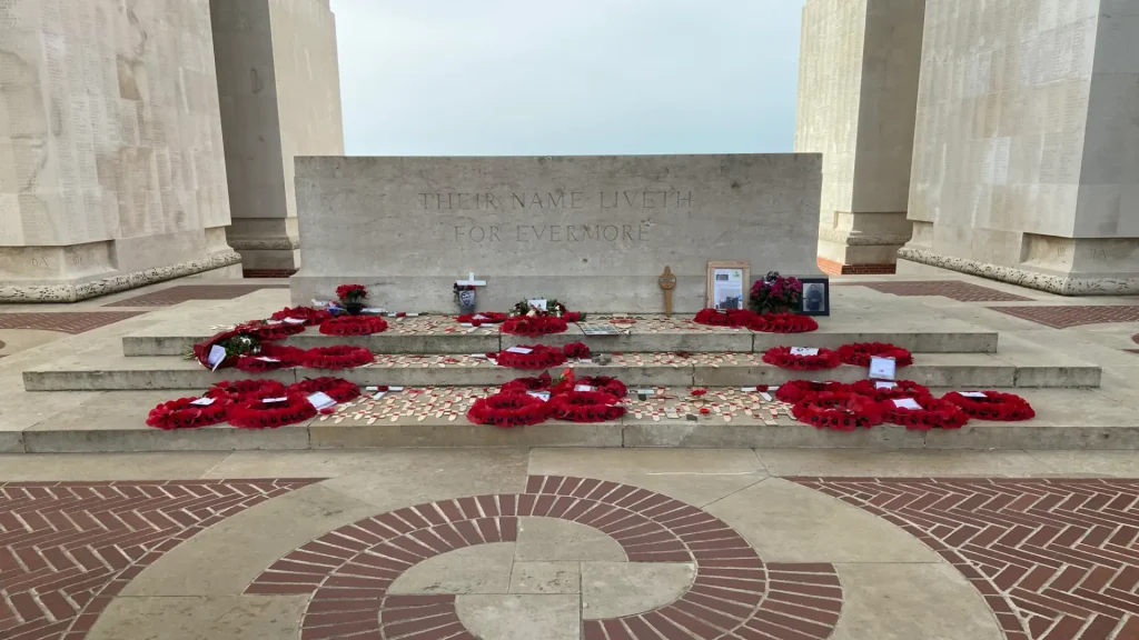 A war memorial with red poppy wreaths lined up on steps, inscribed with "their name liveth for evermore." flanked by stone pillars and a geometric brick floor pattern.