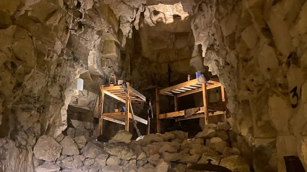 Interior of a historic mine featuring rough stone walls, wooden support beams, and mining tools displayed on platforms illuminated by soft lighting.