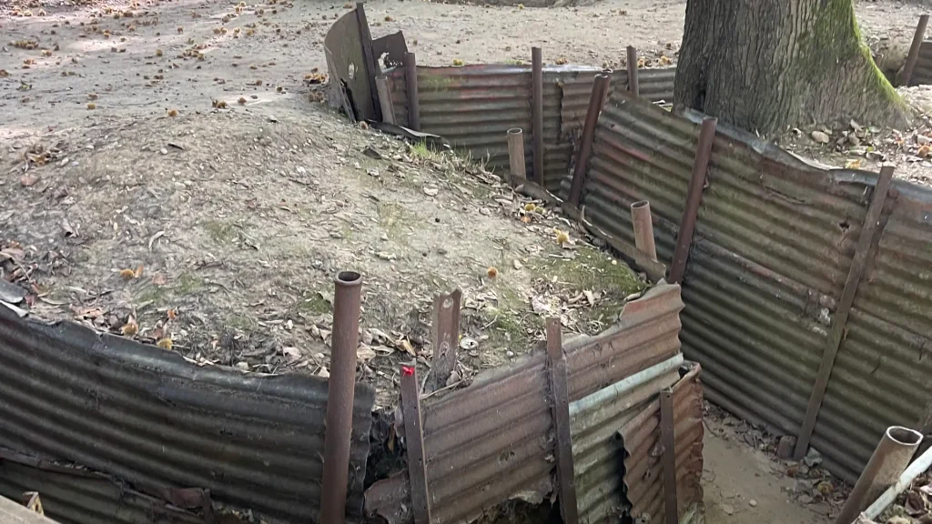 Rusty, corrugated iron sheets forming a trench with exposed pipes set against a backdrop of scattered leaves and bare trees, suggesting a historical or military simulation setting.