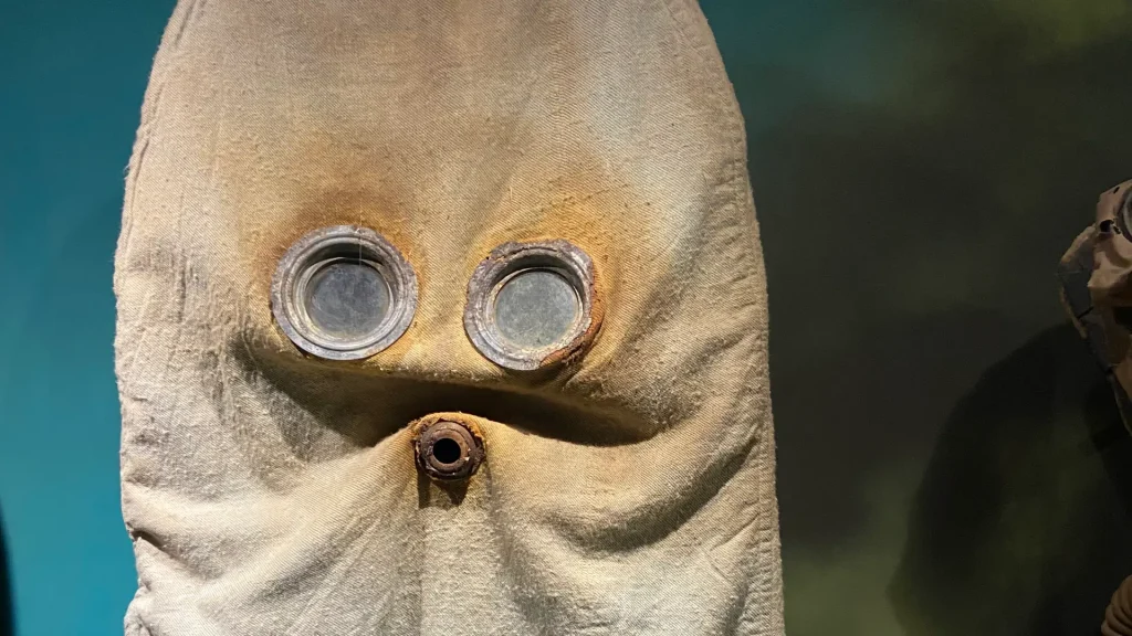 Close-up of a rough textured fabric mask with two large, circular eye holes and a smaller mouth hole, resembling an abstract face. the colors range from beige to brown with shadowy surroundings.