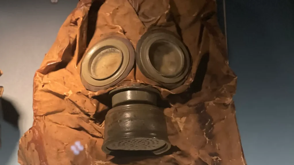 Close-up of a vintage gas mask with large round eye pieces, set against a wrinkled, brown protective hood. the mask is displayed against a dark background.