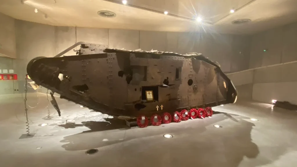 A space capsule displayed in a museum, featuring a charred exterior and numerous heat shield tiles, illuminated under bright lights, with a row of red wreaths placed in front.