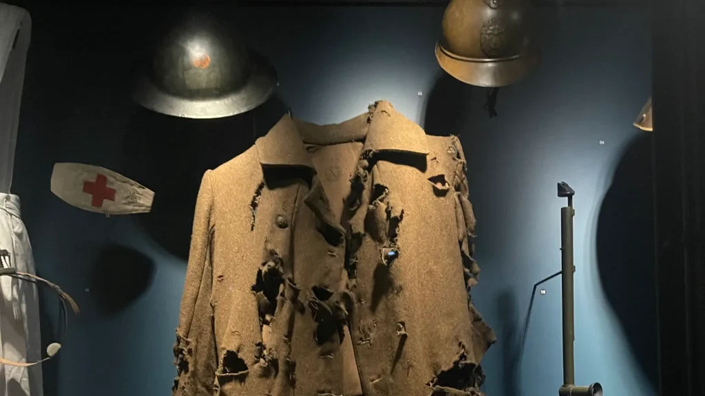 A worn and tattered military uniform with multiple holes displayed in a dimly lit museum exhibit, surrounded by other wartime artifacts including helmets and a medical kit.
