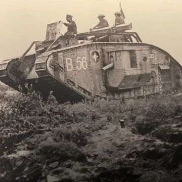 A historic black and white photo of several soldiers riding and standing beside an old military tank moving through a rough terrain field.